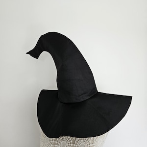 May include: A solid black witch hat featuring a tall, pointed, slightly curved crown and a wide, circular brim. The hat is constructed from a smooth, opaque fabric and is displayed on a white, textured, lace-patterned mannequin form.