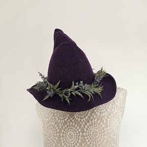 May include: A deep purple knitted pointed hat featuring a wide brim. The base of the crown is adorned with a decorative wreath of artificial green leaves and small light purple lavender-like floral sprigs encircling the entire circumference.