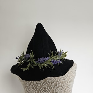 Peut inclure: Un chapeau de sorcière noir à large bord, orné d'une guirlande de fleurs violettes et de feuilles vertes. Le chapeau est présenté sur un mannequin avec un haut en dentelle crème, créant un look fantaisiste et décoratif.
