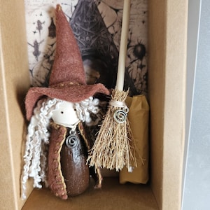 May include: A small wooden doll dressed in a brown felt hat and cape with white curly yarn hair, featuring a silver spiral pendant. Beside the doll is a miniature straw broom with a matching silver spiral charm, all displayed inside a brown box.