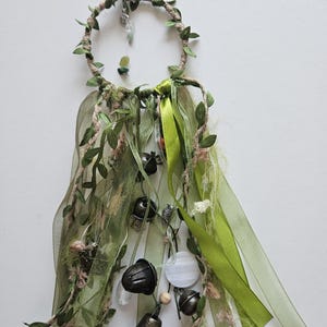 May include: A circular wall hanging featuring a hoop wrapped in twine and artificial green leaves. Long ribbons in shades of olive and lime green hang from the bottom, interspersed with metal bells, small clear crystals, and wooden beads.
