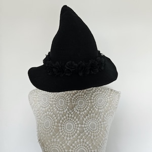 May include: A black pointed hat with a wide brim, featuring a band of small, layered black fabric flowers encircling the base of the crown. The hat has a textured, woven appearance and is displayed on a white, intricate lace-patterned mannequin.