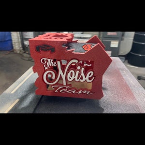 May include: Red wooden box with a cutout design. The box has the words "The Noise Team" written in white on the front.