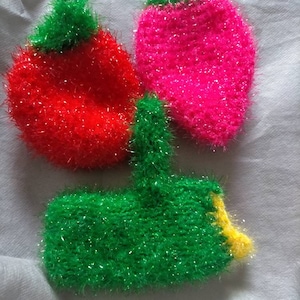 Op de afbeelding: Drie handgemaakte, getextureerde scrubbers in de vorm van fruit. Eén is rood met een groene bovenkant, een andere is felroze met een groene bovenkant en de derde is groen met een gele accent.