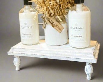 May include: White wooden tiered stand with two clear glass bottles of hand soap labeled "Fir & Leather" with a black pump dispenser. The bottles are sitting on the top tier of the stand. A white ceramic jar with a brown tag that says "Harvest" is sitting on the middle tier of the stand. The jar is filled with dried wheat stalks.