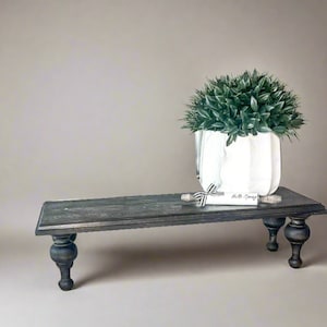Pu&ograve; includere: Un supporto per piante in legno grigio scuro con gambe tornite sostiene una fioriera bianca strutturata piena di fogliame verde. Una piccola pergamena con "Hello Spring" &egrave; legata con un nastro bianco e nero.