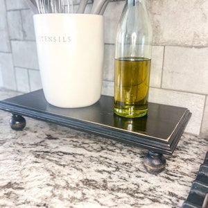 May include: A black wooden tiered stand with turned legs, holding a white ceramic utensil holder with the word "UTENSILS" on it and a clear glass bottle filled with olive oil.