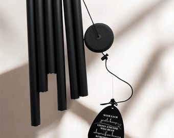 May include: Black wind chimes with a circular top and multiple cylindrical tubes hanging below. A black tag with white text is suspended from the chimes, reading "WITH EACH gentle breeze LISTEN AND YOU WILL HEAR whispers from heaven REMINDING YOU THAT I AM near."