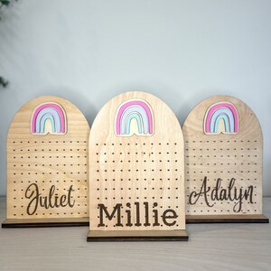 May include: Three wooden earring holders with rainbow cutouts and personalized names. The names are "Juliet", "Millie", and "Adalyn".