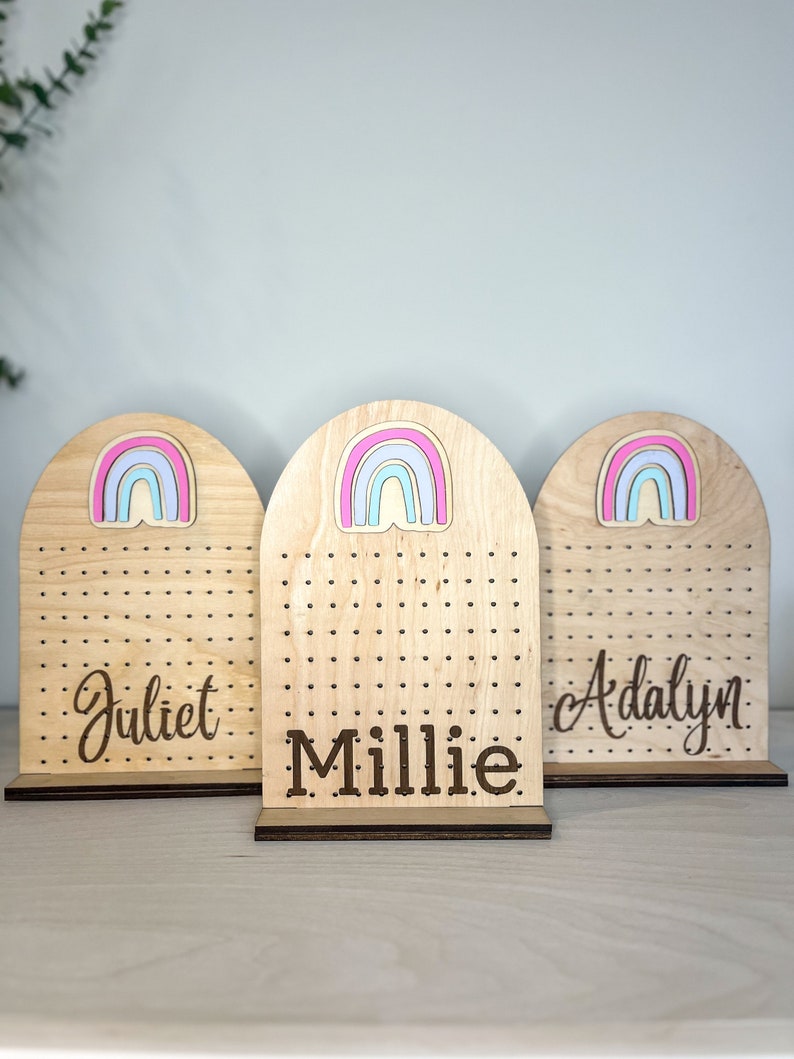 May include: Three wooden earring holders with rainbow cutouts and personalized names. The names are "Juliet", "Millie", and "Adalyn".