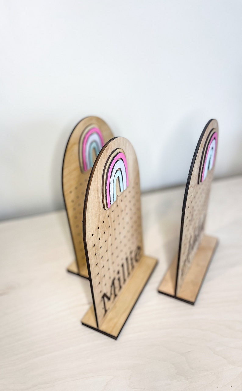 May include: Three wooden earring stands with rainbow-colored accents. The stands are personalized with the names "Millie" and "Jenna".