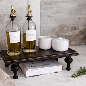 May include: Two glass oil bottles with gold spouts and white labels sit on a dark wooden tray with turned legs. A small white creamer and sugar bowl are also on the tray. A sprig of rosemary rests nearby.
