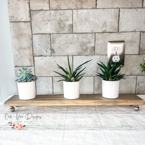 May include: Three potted succulents on a wooden shelf with a white background. The shelf has a distressed wood finish and is supported by two small wooden legs. The text "Oak + Vine Designs HOME DECOR" is visible on the shelf.