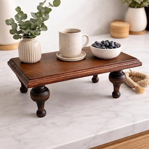 Pu&ograve; includere: Un vassoio di legno marrone scuro con gambe tornite. Il vassoio contiene una tazza con un piattino, una piccola ciotola bianca di mirtilli e un piccolo vaso con del verde. Una corda decorativa &egrave; sul lato.