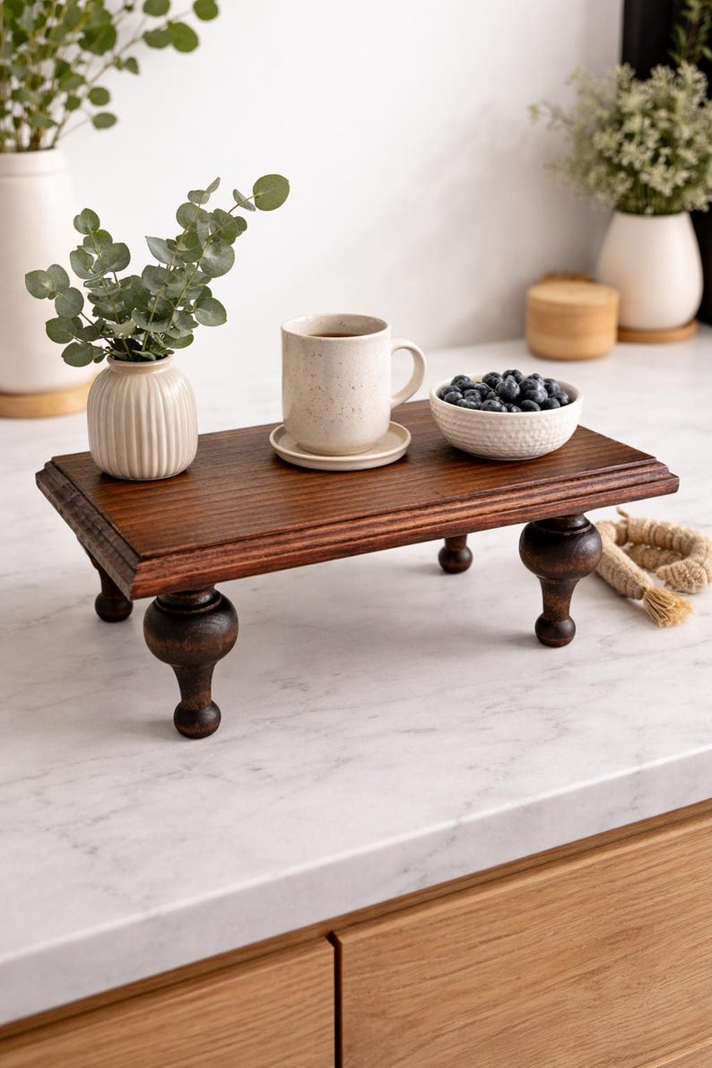 Pu&ograve; includere: Un vassoio di legno marrone scuro con gambe tornite. Il vassoio contiene una tazza con un piattino, una piccola ciotola bianca di mirtilli e un piccolo vaso con del verde. Una corda decorativa &egrave; sul lato.
