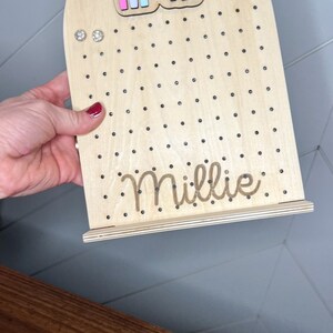 May include: Wooden earring holder with a rainbow design and the name Millie engraved on the bottom. The holder has a stand and is decorated with small black dots.