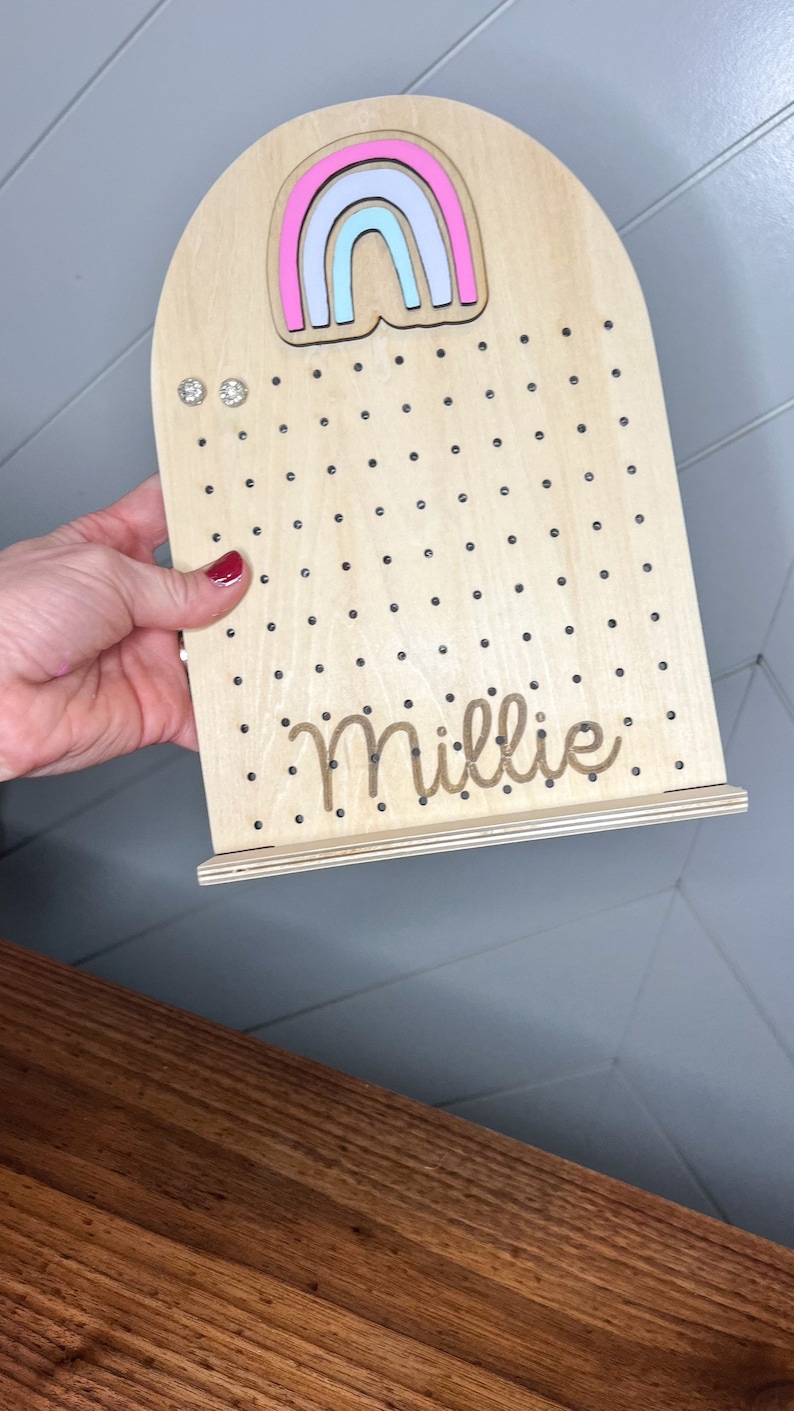 May include: Wooden earring holder with a rainbow design and the name Millie engraved on the bottom. The holder has a stand and is decorated with small black dots.
