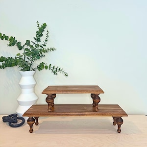 Pu&ograve; includere: Due alzatine in legno con finitura marrone scuro. Le alzatine hanno un piano rettangolare e gambe tornite. Una alzatina &egrave; impilata sull'altra. Lo sfondo include un vaso bianco con vegetazione.