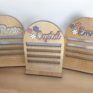 May include: Three wooden earring display stands with names "Rosie", "Crystal", and "Emmy" in pink and purple lettering. Each stand has a floral design with white, pink, and purple flowers.
