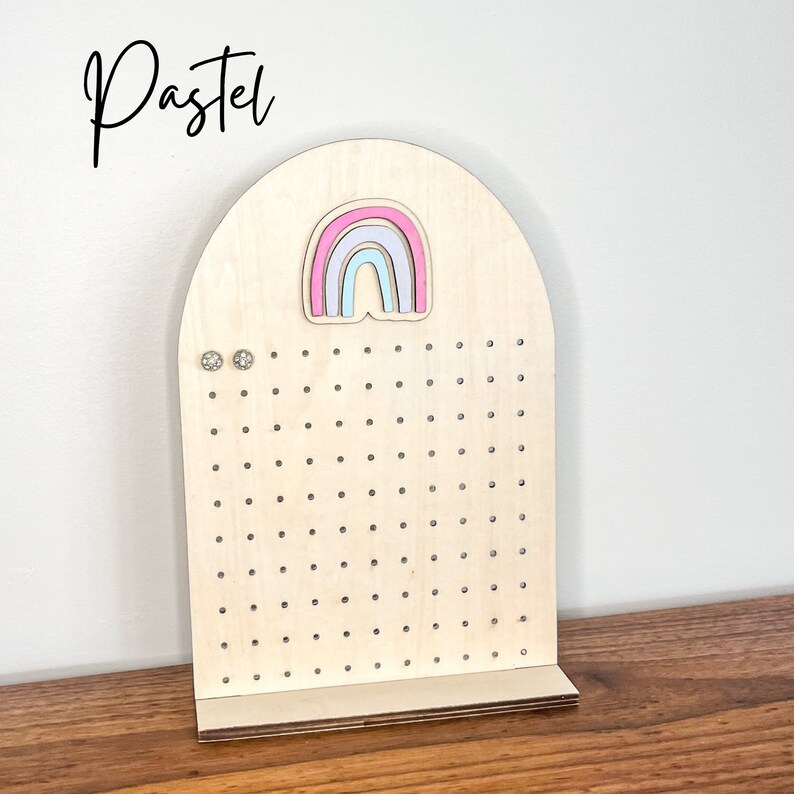 May include: Wooden earring display stand with a rainbow cutout and holes for earrings. The stand is light brown and has a rounded top.
