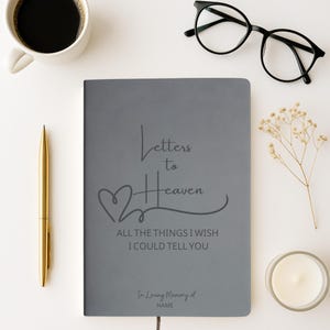 Peut inclure: Un journal gris avec le texte "Letters to Heaven" et "All the things I wish I could tell you." Un stylo doré, une tasse de café, une paire de lunettes noires, une bougie et des fleurs séchées sont disposés autour du journal.