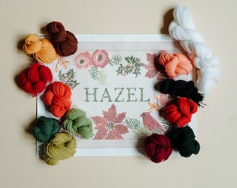 May include: A needlepoint canvas with the name "HAZEL" embroidered in red, surrounded by floral and bird motifs. The canvas is surrounded by various colors of embroidery floss, including red, green, white, and orange.