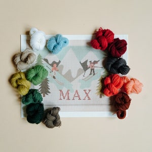 May include: A white canvas with the name "MAX" printed on it, surrounded by 12 balls of yarn in various colours, including red, green, blue, and brown.