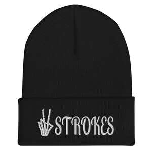 May include: Black beanie with a white embroidered skeleton hand making a peace sign and the word "STROKES" below it.