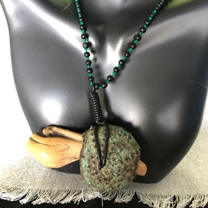 Peut inclure: Un collier artisanal avec un pendentif texturé vert et marron, orné d'un fil de fer et d'un morceau de bois flotté. Le collier est composé de perles noires enfilées sur un cordon vert. Le pendentif est la pièce maîtresse.