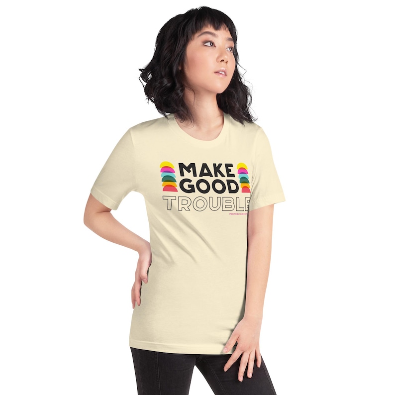 Pode incluir: Uma camiseta de cor creme com mangas curtas e gola redonda. A camiseta tem uma estampa gr&aacute;fica com o texto "MAKE GOOD TROUBLE" em preto e um design colorido de semic&iacute;rculos sobrepostos.