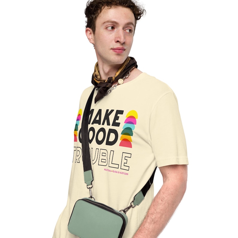Pode incluir: Uma pessoa vestindo uma camiseta amarelo claro com o texto "MAKE GOOD TRUBLE" em letras pretas. A camiseta tem um design gr&aacute;fico colorido com semic&iacute;rculos. A pessoa est&aacute; usando uma bandana preta e marrom ao redor do pesco&ccedil;o e uma bolsa transversal preta com uma bolsa verde claro.