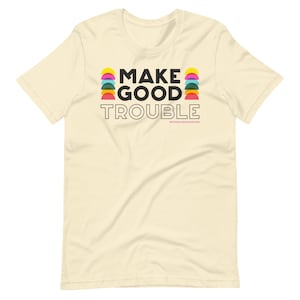 Pode incluir: Uma camiseta bege com o texto "MAKE GOOD TROUBLE" em letras pretas. O texto &eacute; cercado por semic&iacute;rculos coloridos.