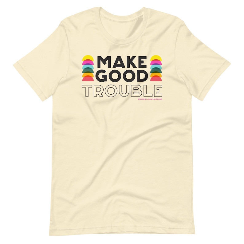 Pode incluir: Uma camiseta bege com o texto "MAKE GOOD TROUBLE" em letras pretas. O texto &eacute; cercado por semic&iacute;rculos coloridos.