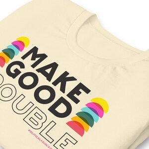 Pode incluir: Uma camiseta bege com o texto "MAKE GOOD DOUBLE" em preto. O texto &eacute; rodeado por um design colorido de semic&iacute;rculos.
