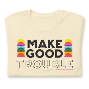 Pode incluir: Uma camiseta cor creme com o texto "MAKE GOOD TROUBLE" em preto. O texto &eacute; cercado por semic&iacute;rculos coloridos. O texto "POLITICALSOULQUEST.COM" est&aacute; em preto menor na parte inferior da camiseta.
