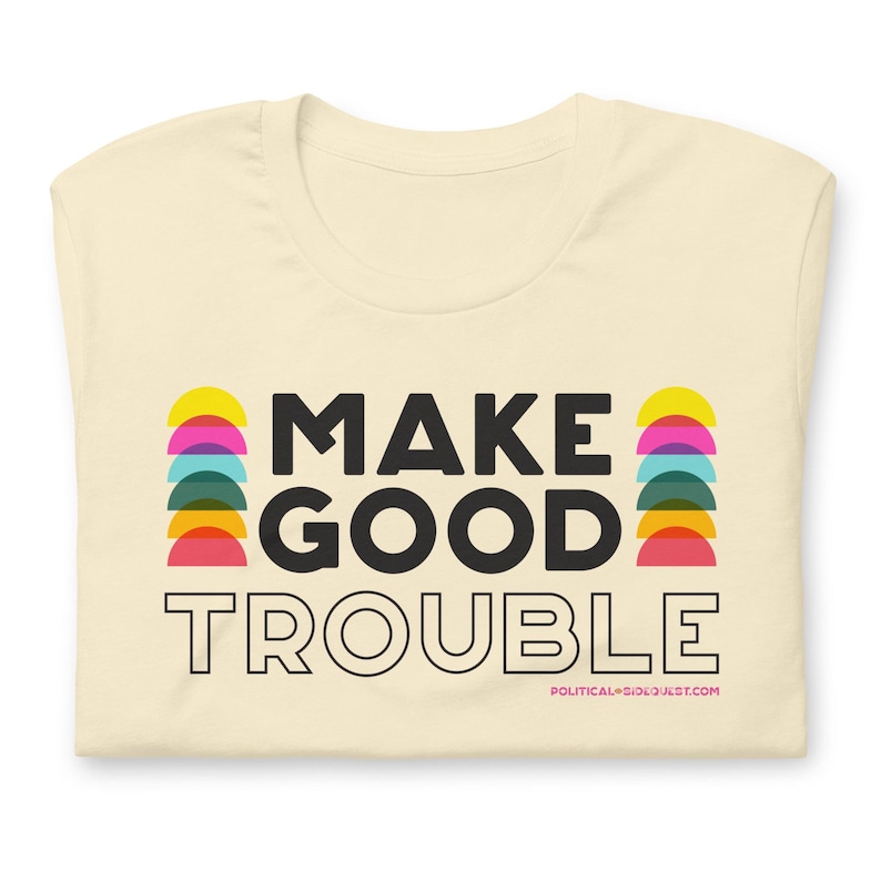 Pode incluir: Uma camiseta cor creme com o texto "MAKE GOOD TROUBLE" em preto. O texto &eacute; cercado por semic&iacute;rculos coloridos. O texto "POLITICALSOULQUEST.COM" est&aacute; em preto menor na parte inferior da camiseta.