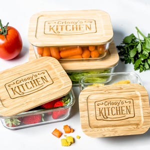 May include: A set of rectangular glass food storage containers with bamboo lids. The lids are engraved with "Crissy's Kitchen". One container holds strawberries, another baby carrots, and a third, lettuce. A tomato and parsley are also visible.
