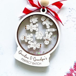 Puede incluir: Adorno navideño de madera blanco con forma de bola de nieve con una cinta roja y blanca. El adorno tiene una cúpula de plástico transparente con purpurina en el interior y recortes de pan de jengibre con nombres escritos en ellos. El adorno dice "Grandma & Grandpa's Perfect Batch" en la parte inferior.