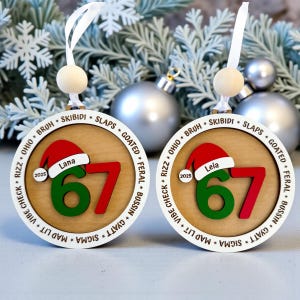 Two Personalized 67 Christmas ornament shown with pine branches and silver Christmas balls. Ornament includes Santa hat with custom name, year, green and red six seven numbers, and border of Gen Z slang words like skibidi, rizz, slaps, goated, feral, bussin, sigma, mad lit, vibe check, and Ohio. Ideal funny Christmas gift for teens, Gen Z, and stocking stuffers.