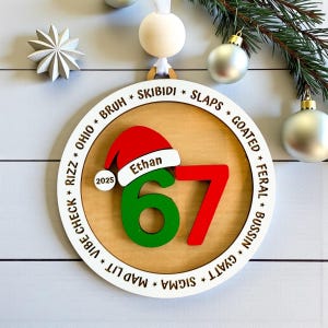 Personalized 67 Christmas ornament shown in a flat lay with pine branches and silver Christmas balls. Ornament includes Santa hat with custom name, year, green and red six seven numbers, and border of Gen Z slang words like skibidi, rizz, slaps, goated, feral, bussin, sigma, mad lit, vibe check, and Ohio. Ideal funny Christmas gift for teens, Gen Z, and stocking stuffers.