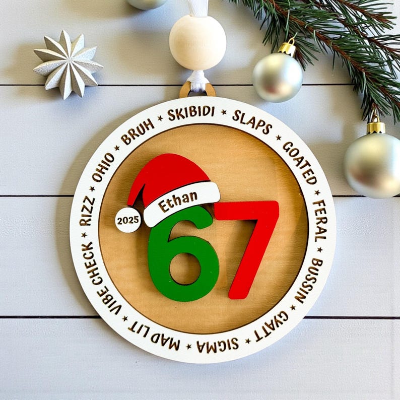 Personalized 67 Christmas ornament shown in a flat lay with pine branches and silver Christmas balls. Ornament includes Santa hat with custom name, year, green and red six seven numbers, and border of Gen Z slang words like skibidi, rizz, slaps, goated, feral, bussin, sigma, mad lit, vibe check, and Ohio. Ideal funny Christmas gift for teens, Gen Z, and stocking stuffers.