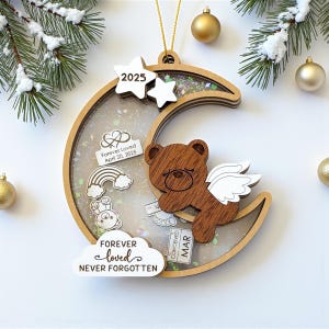 Puede incluir: Un adorno navideño de madera con forma de media luna con un centro transparente lleno de purpurina y abalorios. Un oso pardo con alas blancas está adjunto. El adorno incluye el texto "2025", "Forever Loved" y "Forever Loved, Never Forgotten".