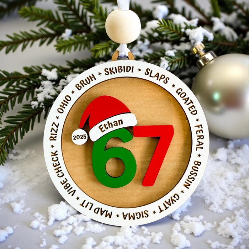 Close-up of custom 67 Christmas ornament with Santa hat, name and year personalization, green and red layered numbers, and a slang-filled border including skibidi, rizz, goated, bussin, slaps, sigma, and vibe check. Ornament is styled with mini Christmas trees and silver ornaments on a frosted tabletop. Perfect personalized Christmas gift for teen boys, teen girls, and Gen Z stocking stuffers.