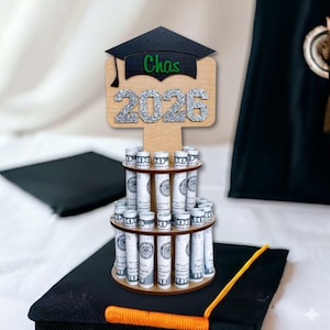 Graduation money tree gift 2026 in natural wood with silver glitter “2026” and personalized name. Wooden graduation money holder displayed on a graduation cap with rolled US dollar bills. Graduation decorations Class of 2026 cash gift for her or him.