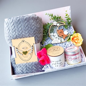 May include: A baby gift basket with a gray plush blanket, a wooden ornament with a bear, and a candle. The basket includes a wooden tag with the text "CARRIED FOR A MOMENT, LOVED FOR A LIFETIME". A candle with the text "no words, just hugs" is also included.