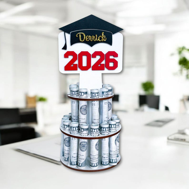 Graduation money holder 2026 stand with bold red “2026” and personalized name on black graduation cap topper. Money tree gift holds rolled US dollar bills for a graduation cash gift. Great graduation decorations Class of 2026 for party table display.