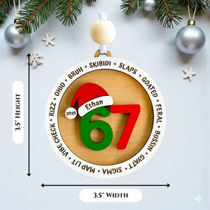 Personalized “67” Gen Z slang Christmas ornament with engraved teen slang words around the border, shown in a flat lay with holiday décor. Ornament features a red and green layered 67 design, Santa hat with custom name, and 2025 year charm. Modern 3.5" x 3.5" round ornament with clean white outer ring and wood-look center. Fun, trendy Christmas gift for teens and tweens, custom stocking stuffer idea.