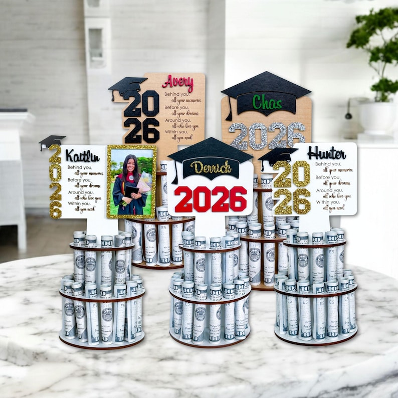 Personalized graduation money holder 2026 collection shown on marble tabletop. Wooden graduation money tree gift with rolled US dollar bills, Class of 2026 designs, graduation cap toppers, and custom names. Graduation decorations Class of 2026 and creative cash gift idea.