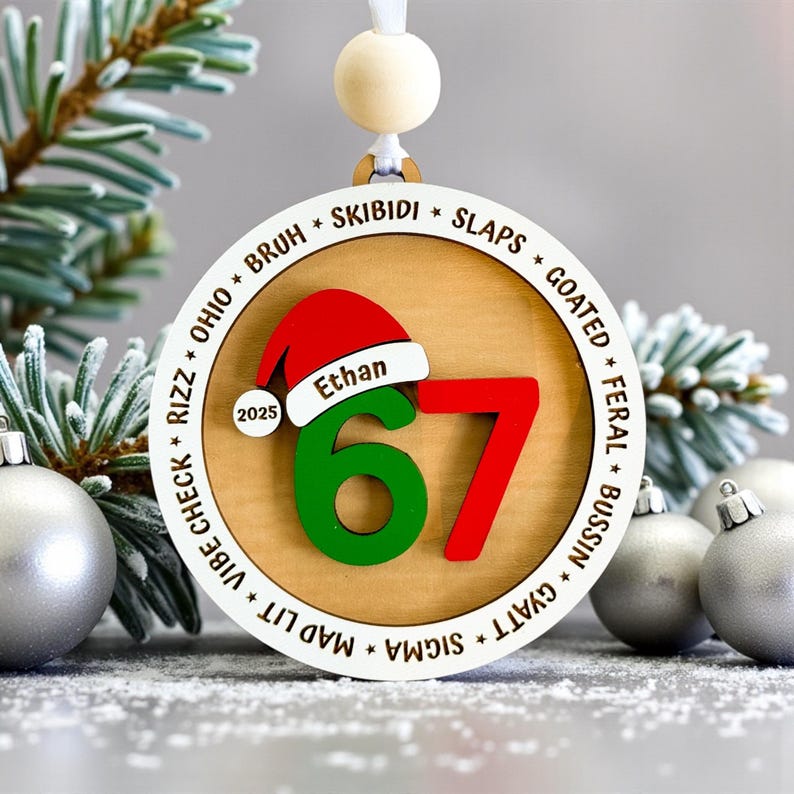 Personalized 67 Christmas ornament shown in a flat lay with pine branches and silver Christmas balls. Ornament includes Santa hat with custom name, year, green and red six seven numbers, and border of Gen Z slang words like skibidi, rizz, slaps, goated, feral, bussin, sigma, mad lit, vibe check, and Ohio. Ideal funny Christmas gift for teens, Gen Z, and stocking stuffers.