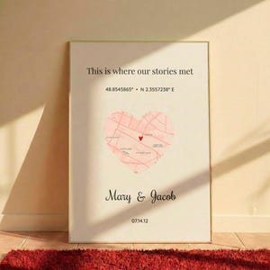 Pode incluir: Impressão emoldurada com o texto "This is where our stories met". Um mapa em forma de coração rosa com um marcador vermelho está centrado na impressão. Os nomes "Mary & Jacob" e a data "07.14.12" estão abaixo do mapa.